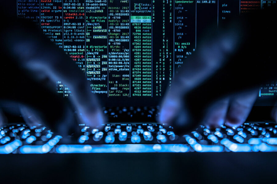 Hände tippen auf leuchtender Tastatur, bunte Codezeilen auf Monitor im Hintergrund.