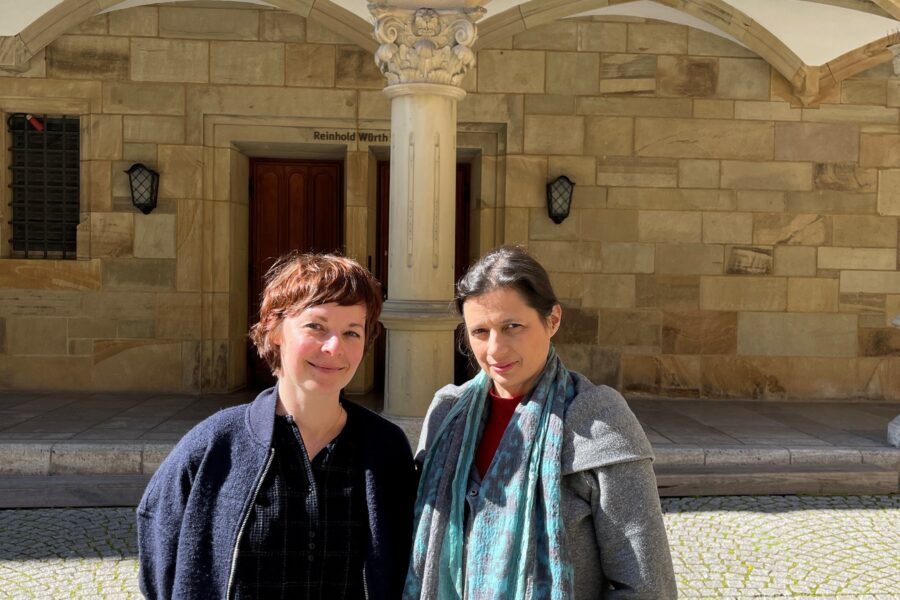 Zwei Personen stehen vor einem Gebäude mit Steinbögen und Balustrade.