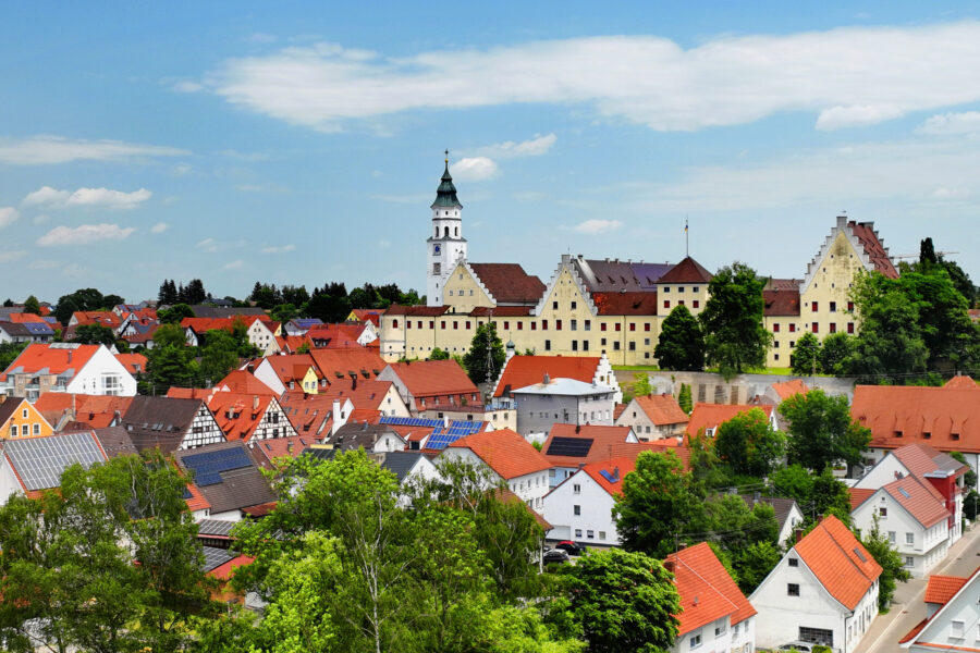Stadtbild mit roten Dächern, gelbem Gebäude und Kirchturm vor blauem Himmel.