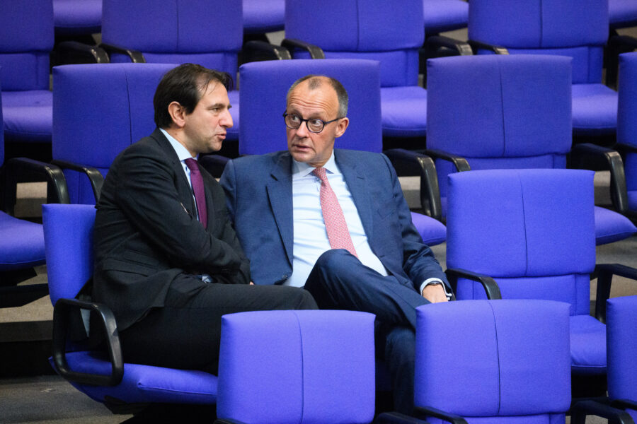 Zwei Männer in Anzügen unterhalten sich in einer Reihe blauer Stühle.