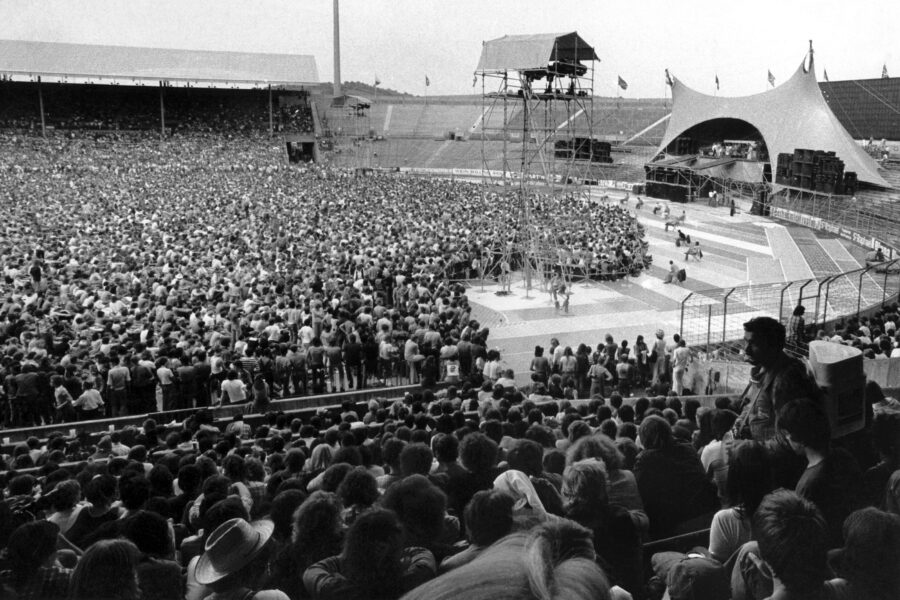 Mehrere Menschen auf Stadiontribünen und vor einer großen Bühne im Freien.