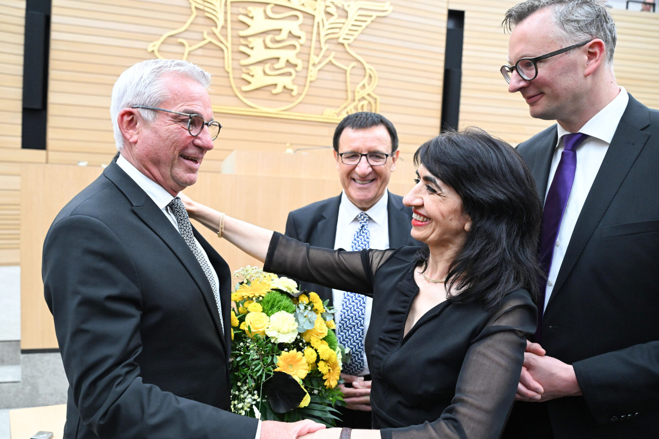 Vier Personen in formeller Kleidung, eine Frau mit Blumenstrauß. Hintergrund: Wappen.