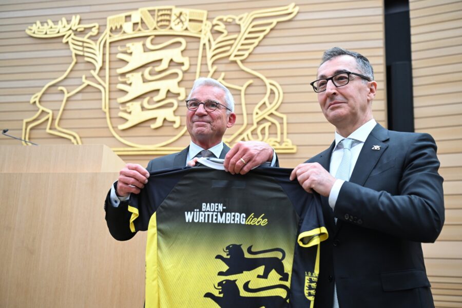 Zwei Männer halten Trikot mit Text "Baden-Württemberg Liebe", im Hintergrund Wappen.