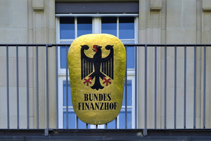 Schild mit Bundesadler und Text "Bundesfinanzhof" auf einem Geländer vor Fenster.