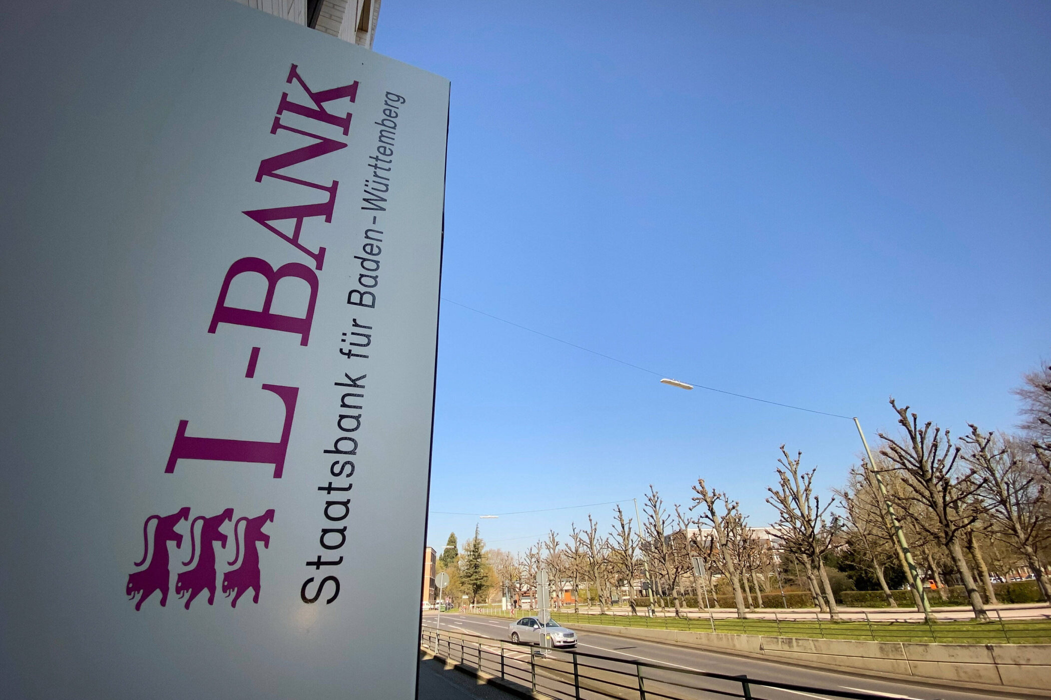 Schild mit Text „L-BANK Staatsbank für Baden-Württemberg“ neben Straße und Bäumen.