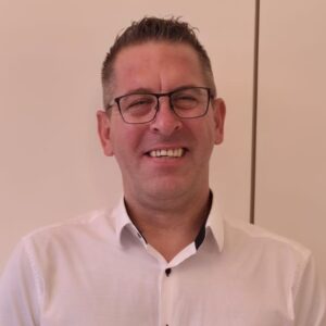 Eine Person mit Brille trägt ein weißes Hemd und lächelt vor einer hellen Wand.