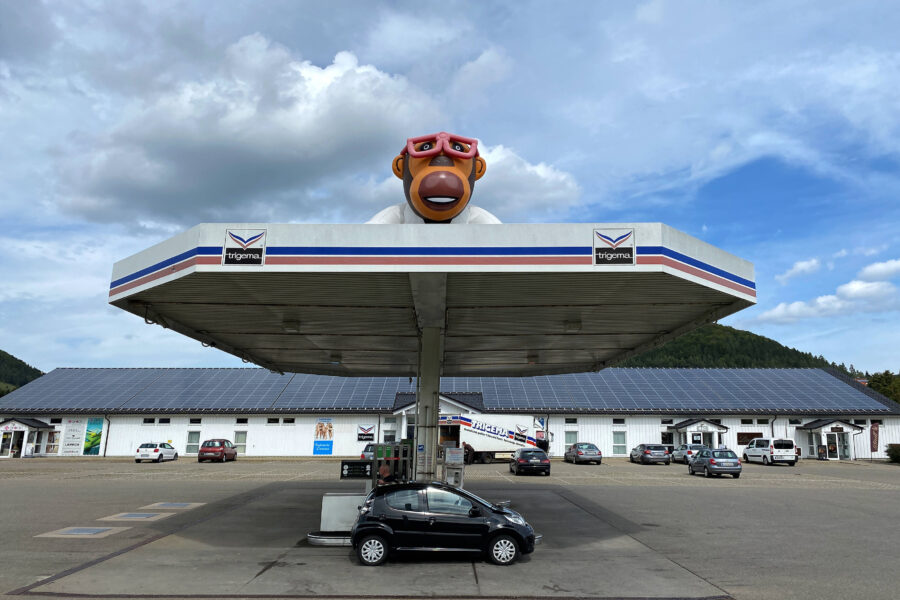 Tankstelle mit Affenskulptur auf Dach, schwarzes Auto an Zapfsäule, Gebäude im Hintergrund.