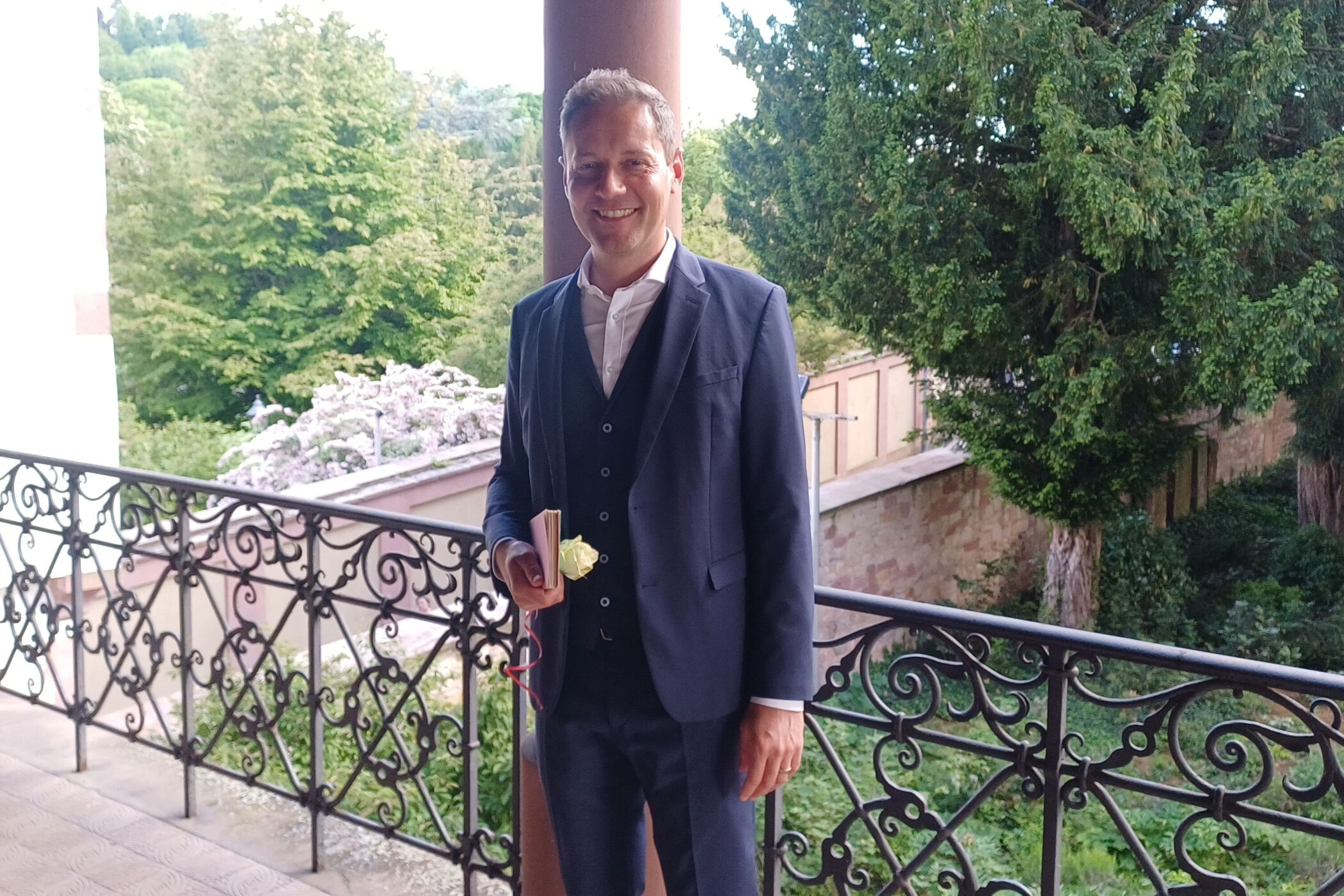 Mann in Anzug steht draußen auf einem Balkon, hält eine gelbe Blume.