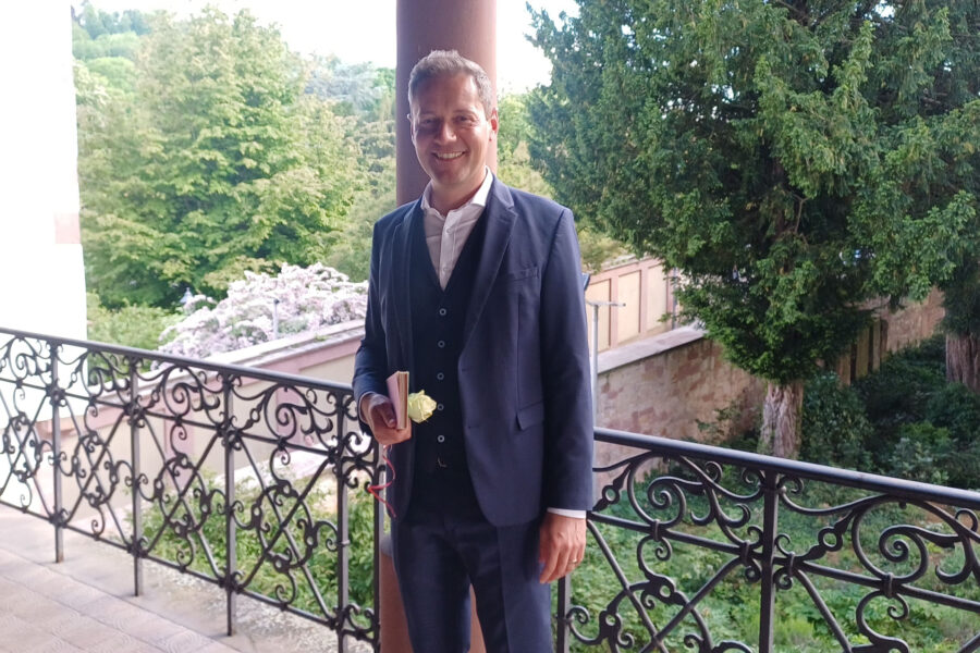 Mann in Anzug steht draußen auf einem Balkon, hält eine gelbe Blume.