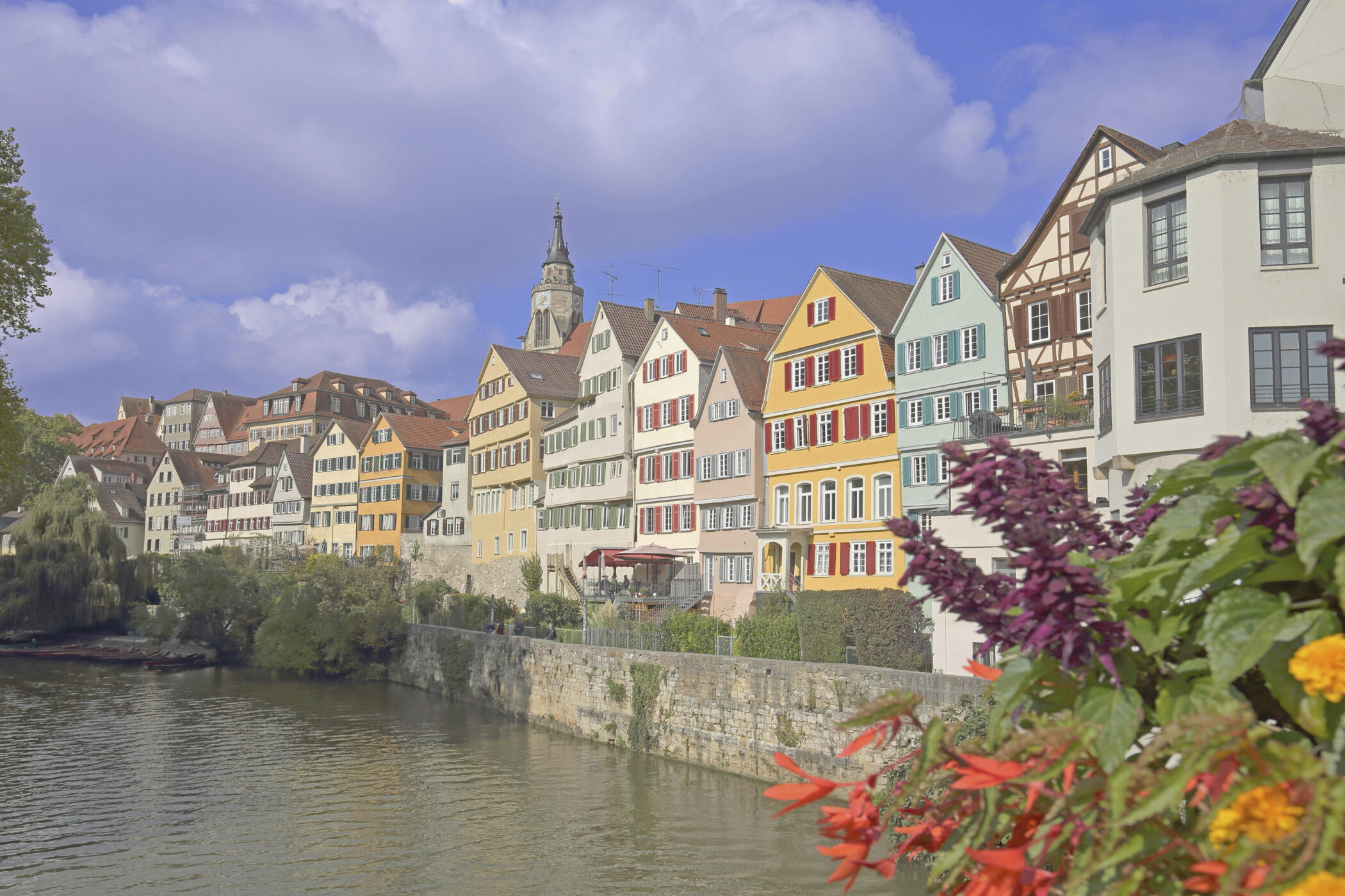 Bunte Fachwerkhäuser am Fluss, Blumen im Vordergrund, Kirchturm im Hintergrund.