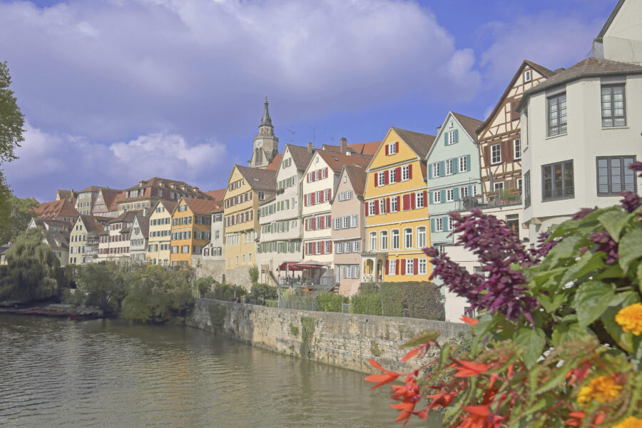 Bunte Fachwerkhäuser am Fluss, Blumen im Vordergrund, Kirchturm im Hintergrund.