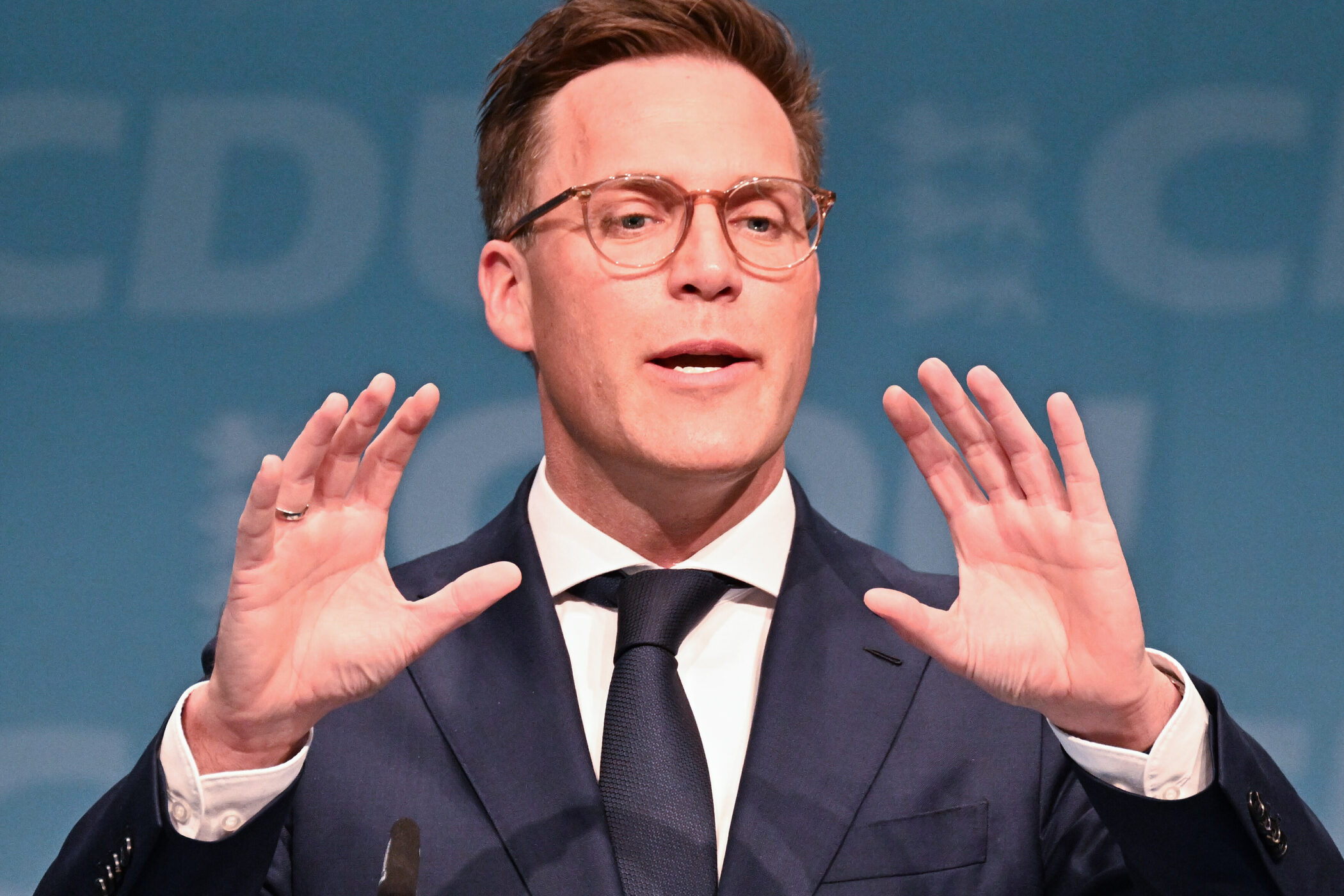 Mann mit Brille im Anzug spricht, hebt Hände. Hintergrund mit verschwommenem Text.