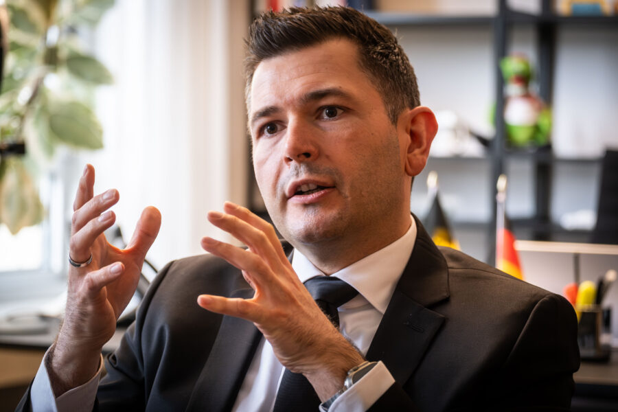 Mann in Anzug gestikuliert in Büro, unscharfer Hintergrund mit Pflanzen und Flaggen.