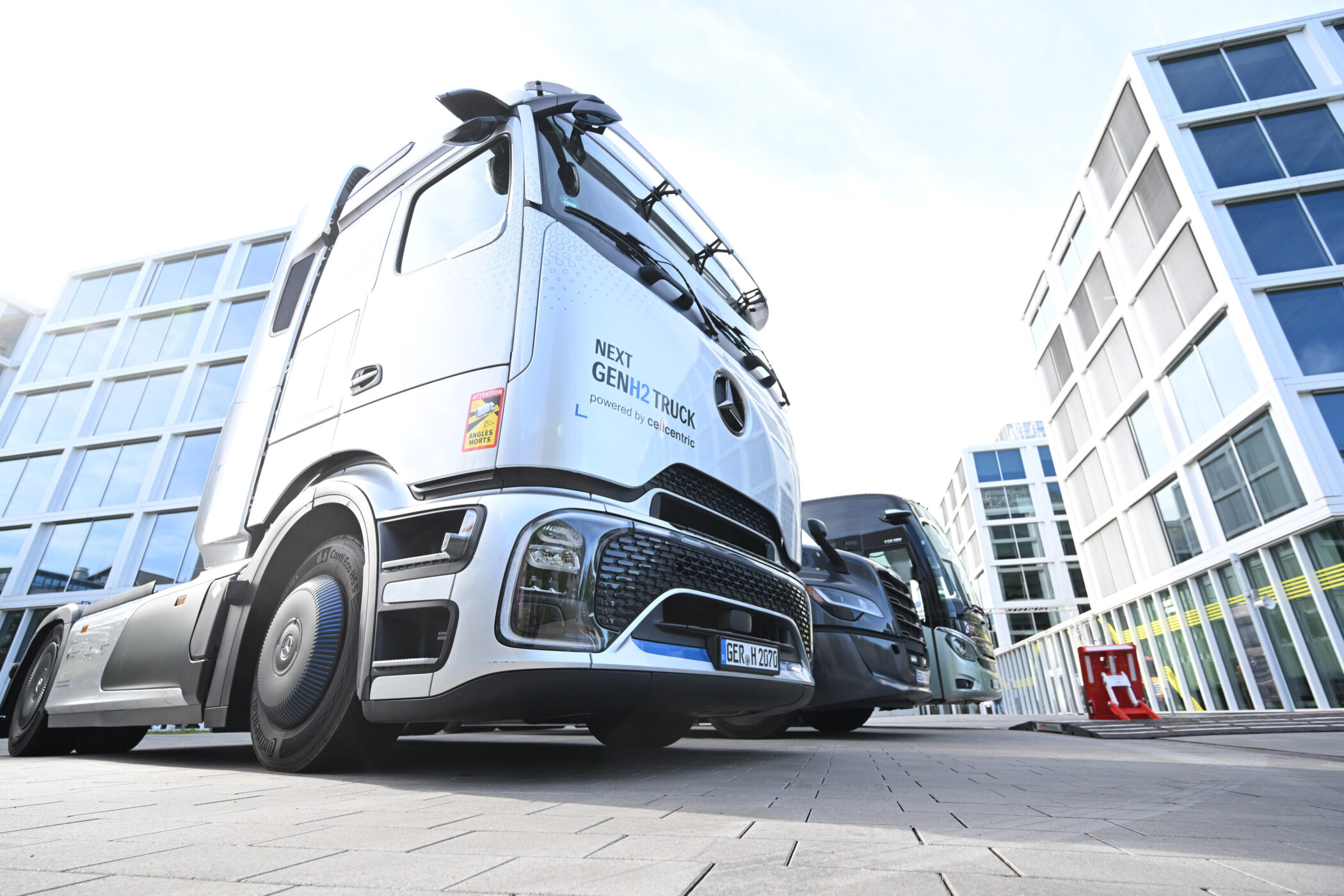 Zwei Lastwagen vor modernen Bürogebäuden, "NEXT GEN H2 TRUCK" auf einem zu lesen.