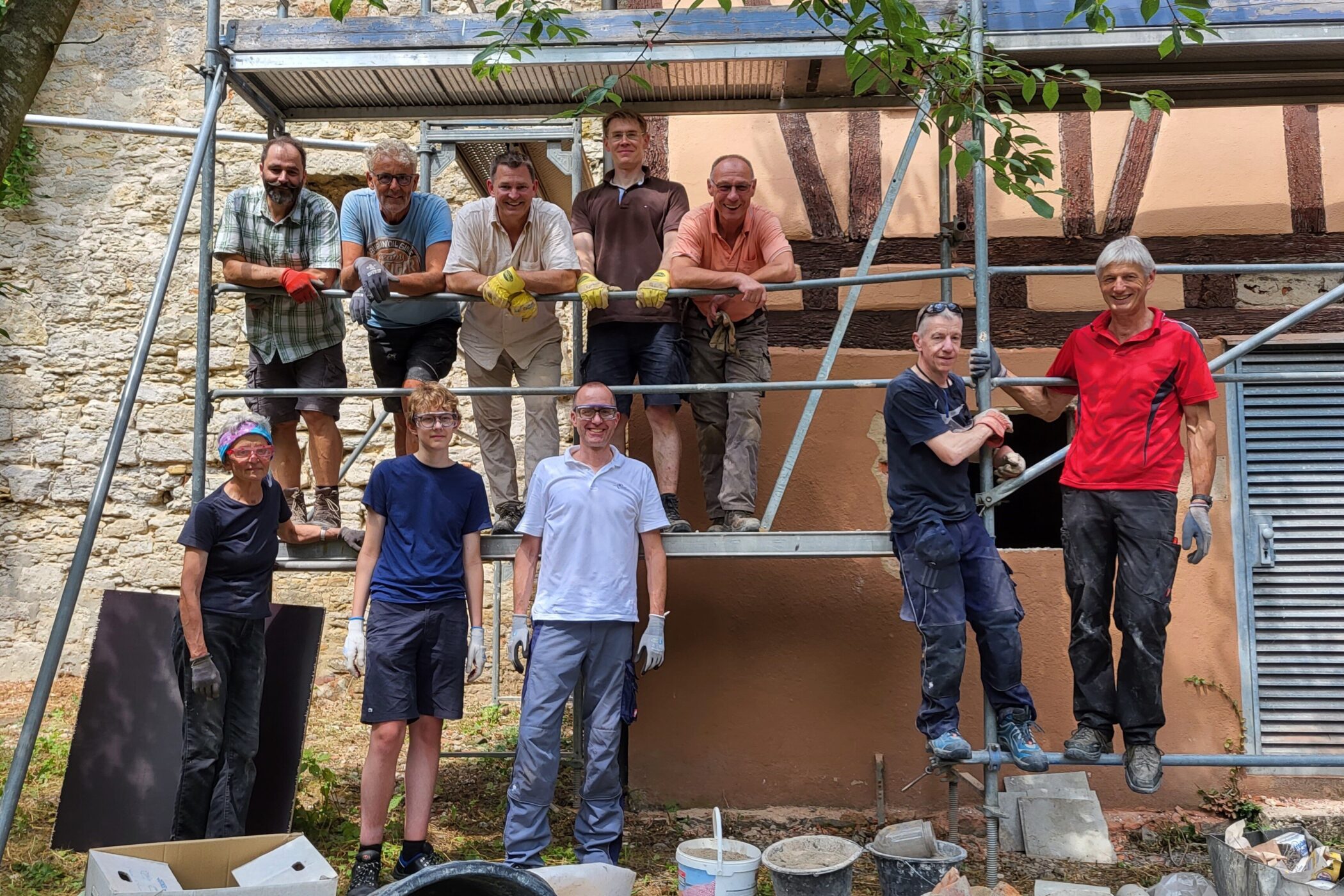 Neun Personen auf einem Baugerüst vor einem Gebäude. Baumaterialien am Boden.