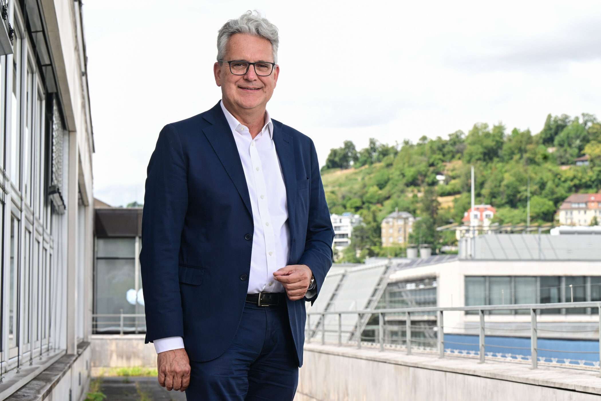 Mann im Anzug steht draußen, Gebäude und Bäume im Hintergrund.