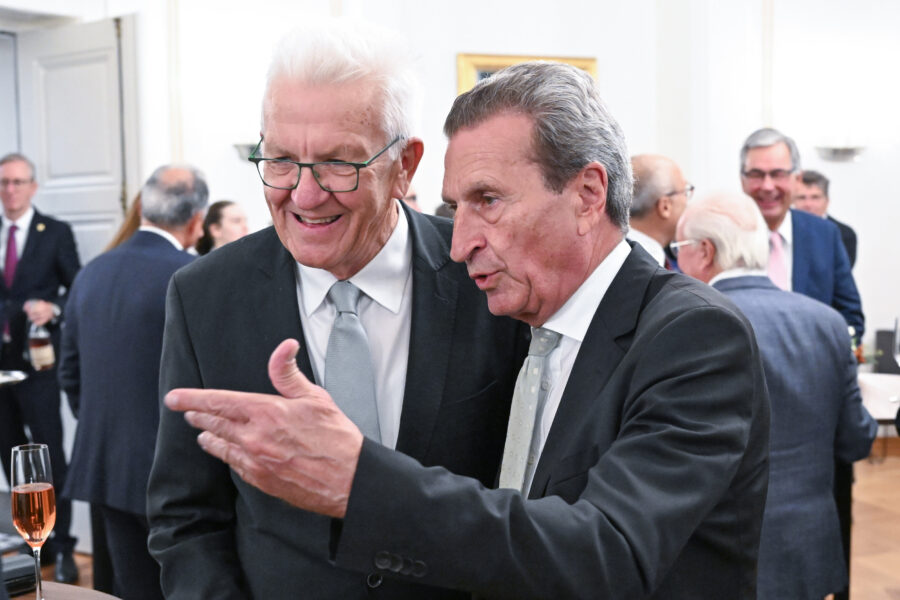 Zwei Männer in Anzügen unterhalten sich in einem Raum mit anderen Personen.