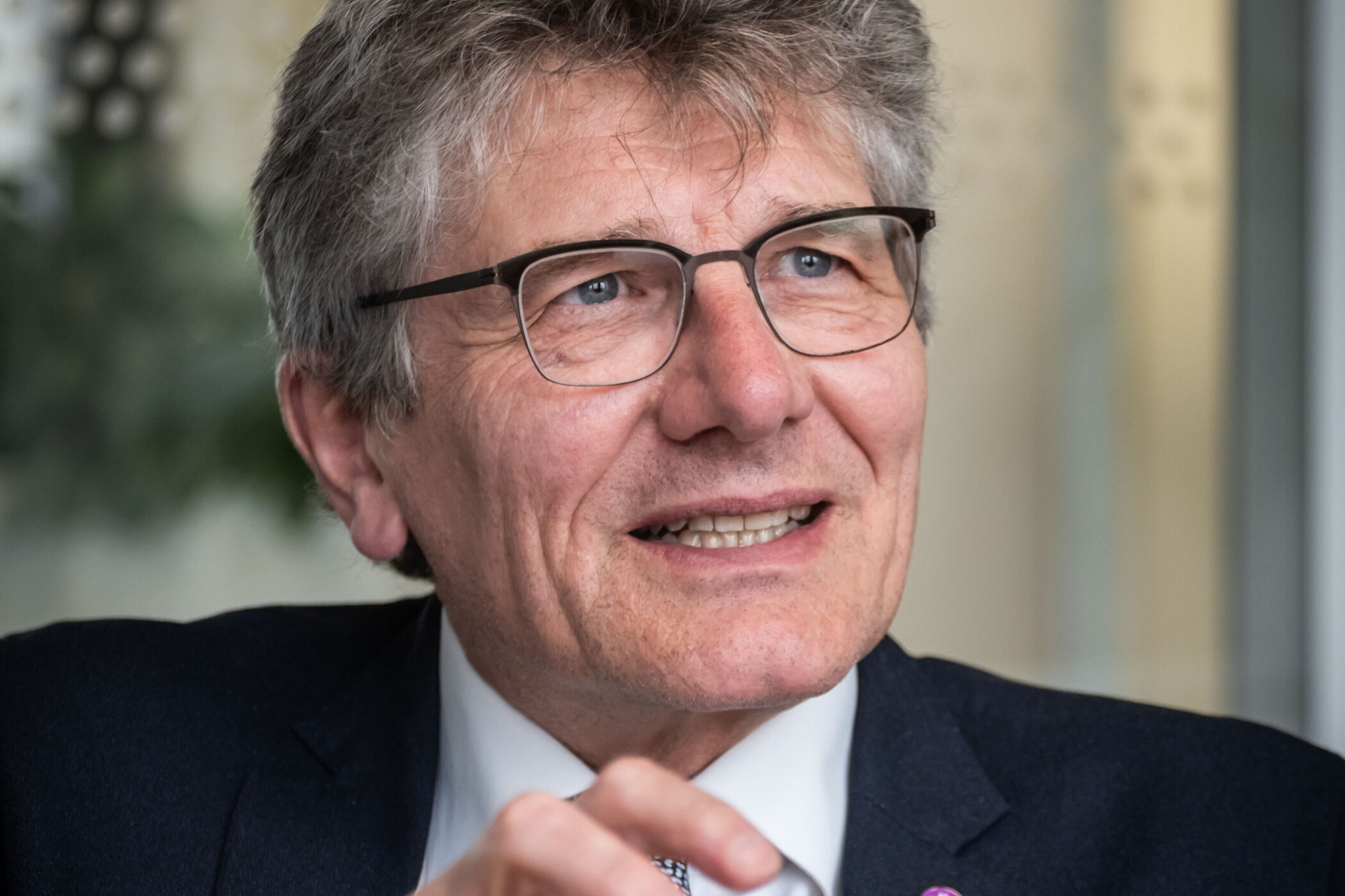 Ein Mann spricht und gestikuliert, Brille und dunkler Anzug mit Anstecknadel.