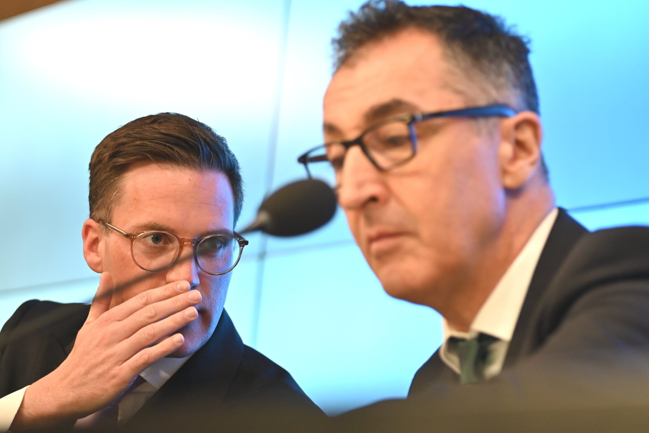 Zwei Männer in Anzügen, einer spricht ins Mikrofon, der andere hält Hand an den Mund.