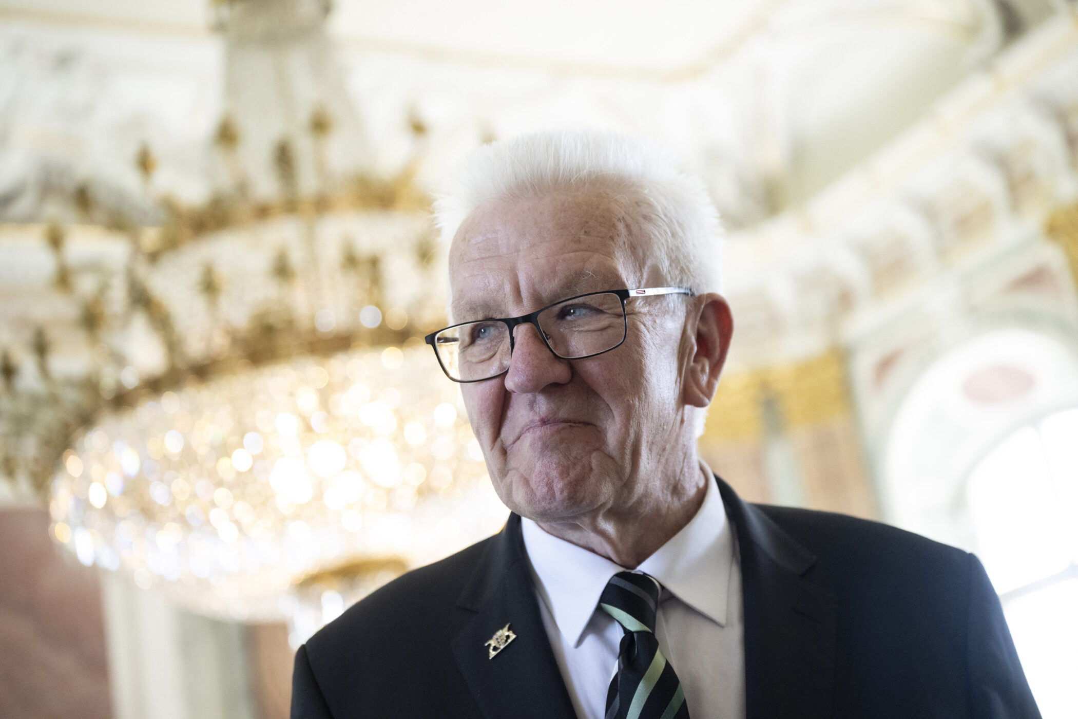 Älterer Mann mit Brille und Anzug vor großem Kronleuchter.