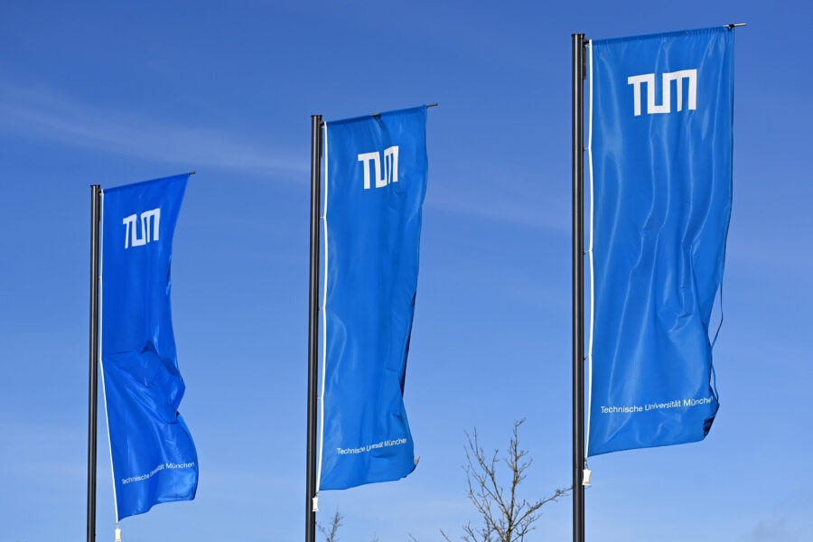 Drei blaue Fahnen mit weißem TUM-Logo vor blauem Himmel.