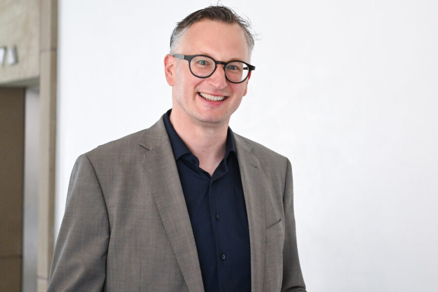 Mann mit Brille in grauem Anzug und dunkelblauem Hemd lächelnd.