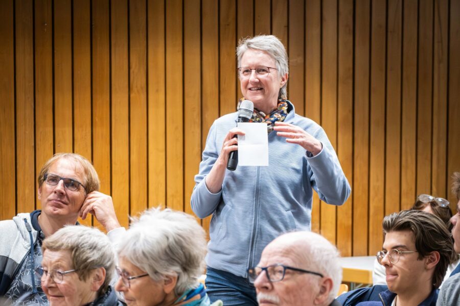 Eine Person spricht in ein Mikrofon vor Publikum, Holzverkleidung im Hintergrund.