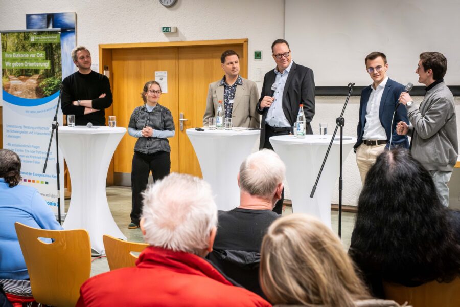 Fünf Personen stehen an runden Tischen, Publikum im Vordergrund.