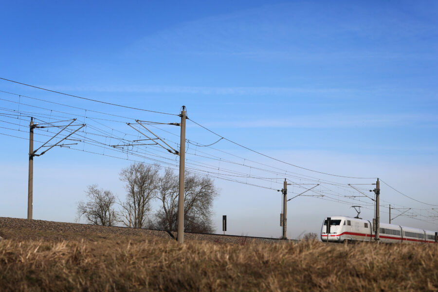 Ein Zug fährt unter Stromleitungen durch eine ländliche Gegend.