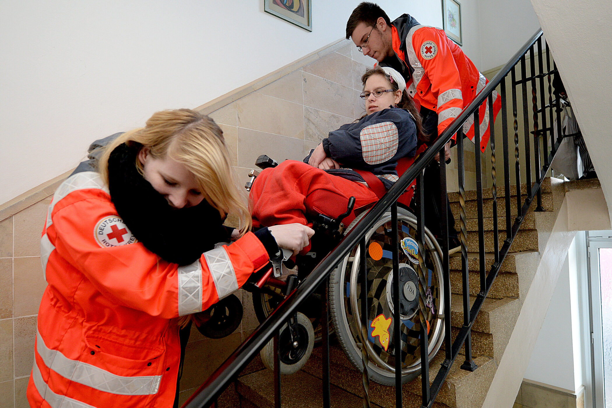 Zwei Personen in roten Jacken helfen einer Person im Rollstuhl die Treppe hinab.
