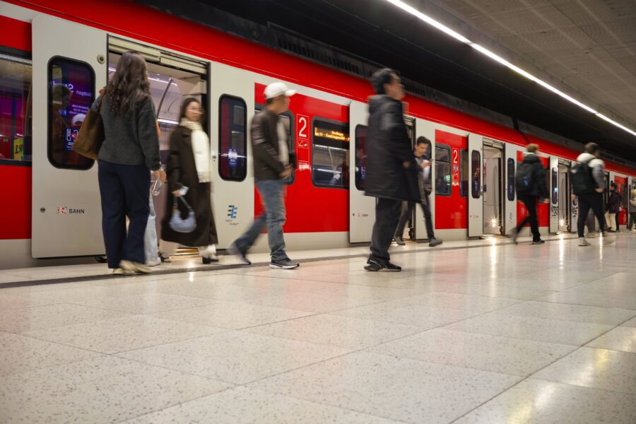 Menschen steigen in einen roten Zug mit offenen Türen auf einem Bahnsteig ein.