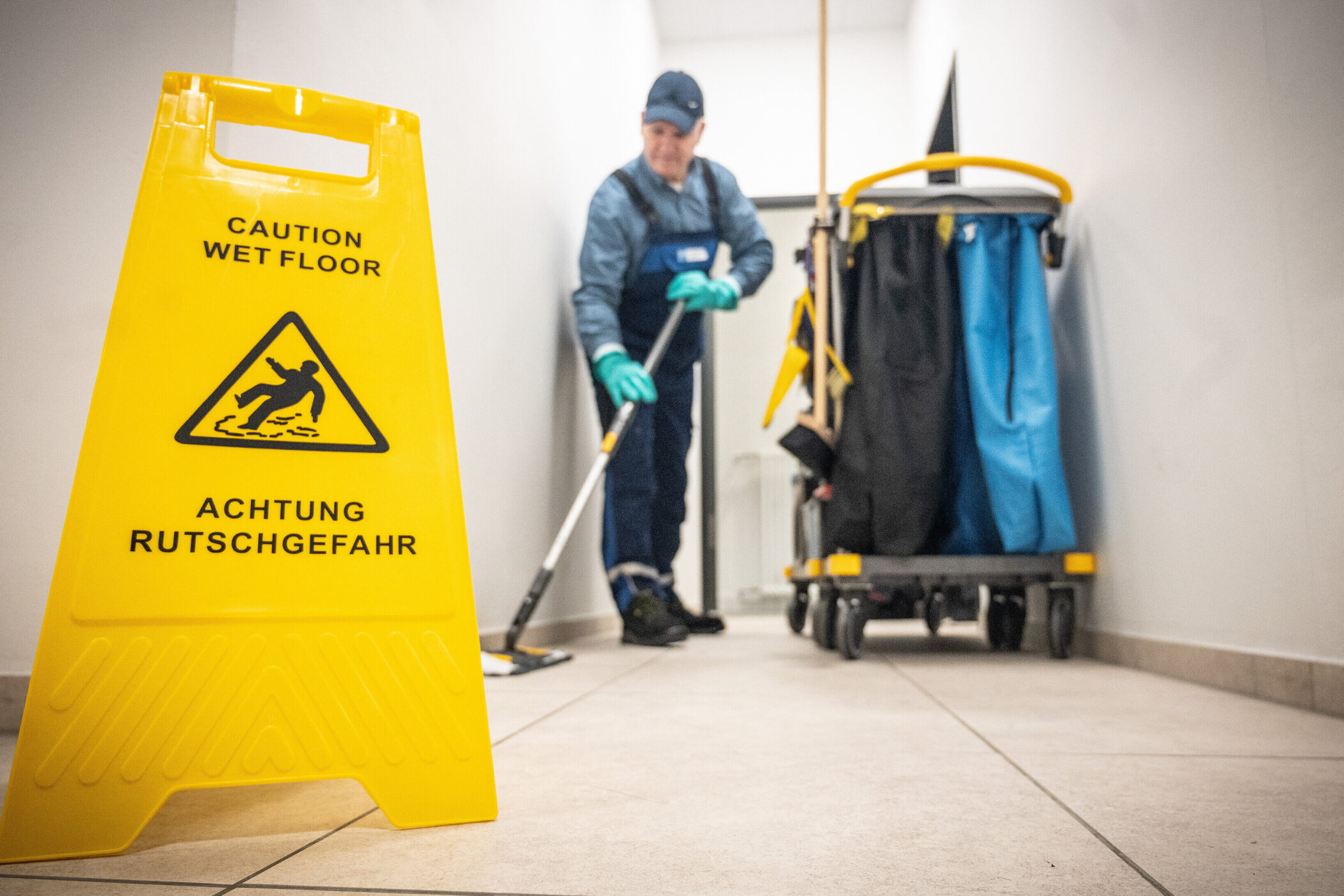 Gelbes Warnschild „Achtung Rutschgefahr“ vor Reinigungskraft mit Putzwagen.