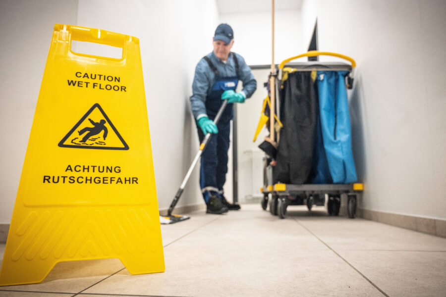 Gelbes Warnschild „Achtung Rutschgefahr“ vor Reinigungskraft mit Putzwagen.