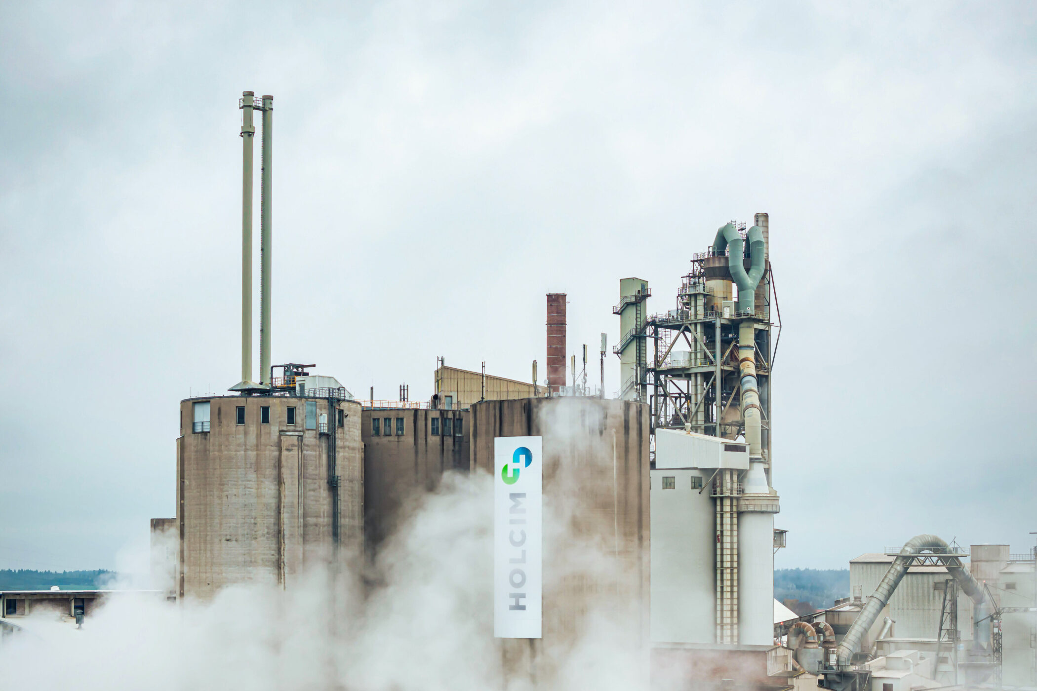 Industrieanlage mit Schornsteinen, Text "HOLCIM" an der Wand, umgeben von Nebel.