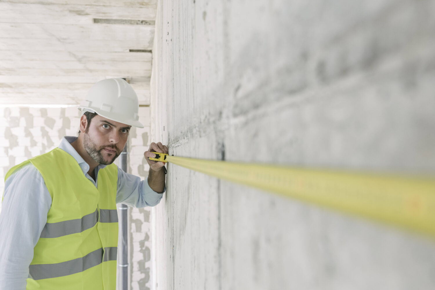 Person mit Helm und Weste misst Betonwand mit einem Maßband.