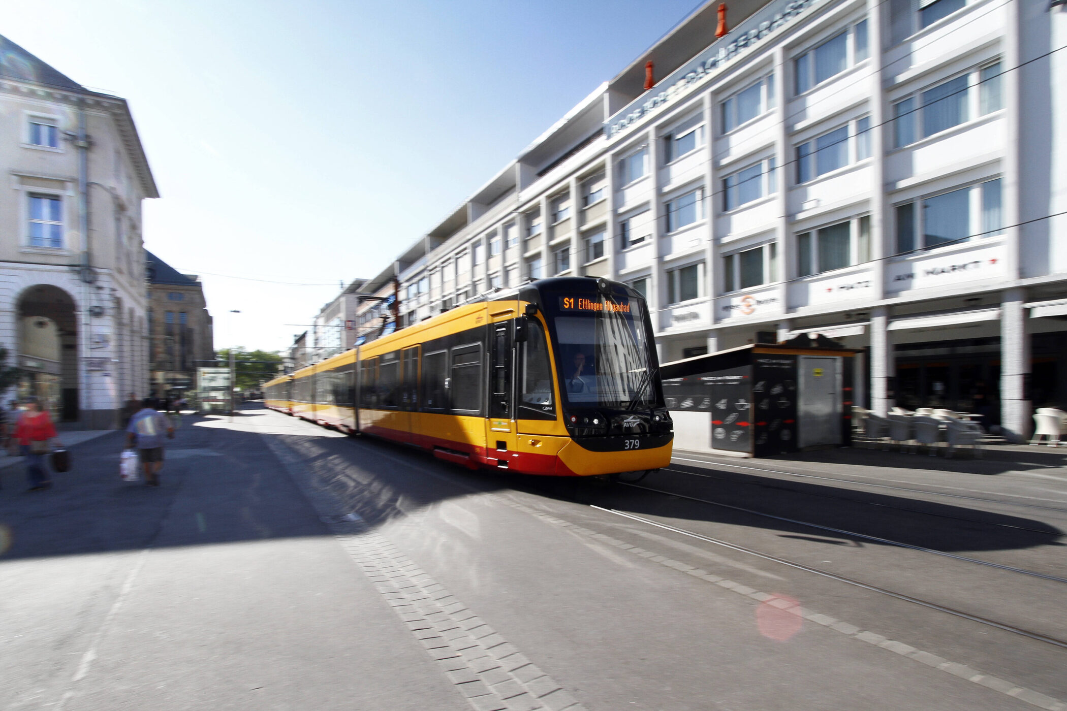Gelb-rote Straßenbahn auf Straße, Gebäudefassade und Fußgänger im Hintergrund.