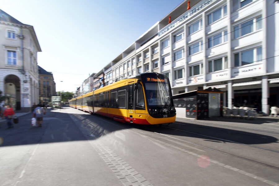 Gelb-rote Straßenbahn auf Straße, Gebäudefassade und Fußgänger im Hintergrund.