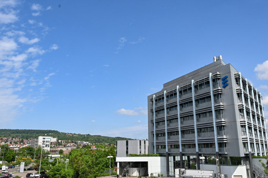 Graues Bürogebäude mit blauem Logo, angrenzende Parkplätze, blauer Himmel.