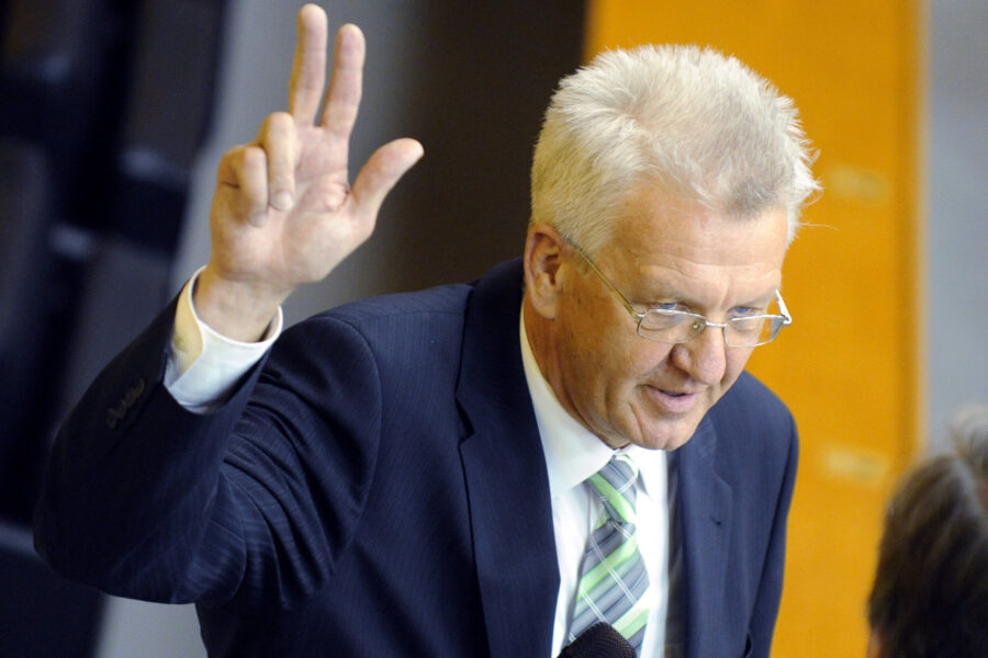 Älterer Mann im Anzug spricht und zeigt eine Handgeste mit drei Fingern.