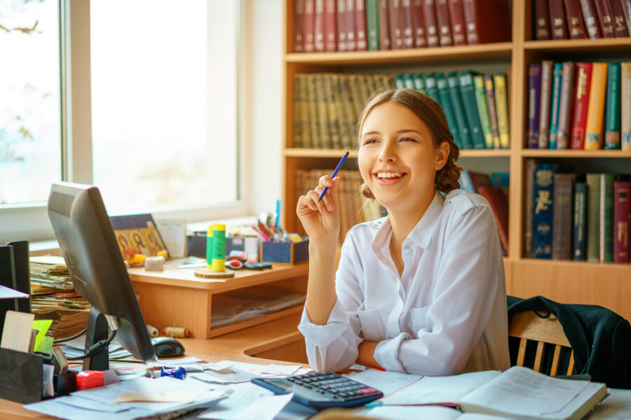 Person am Schreibtisch mit Computer, Büchern, Ordnern und einem Stift in der Hand.