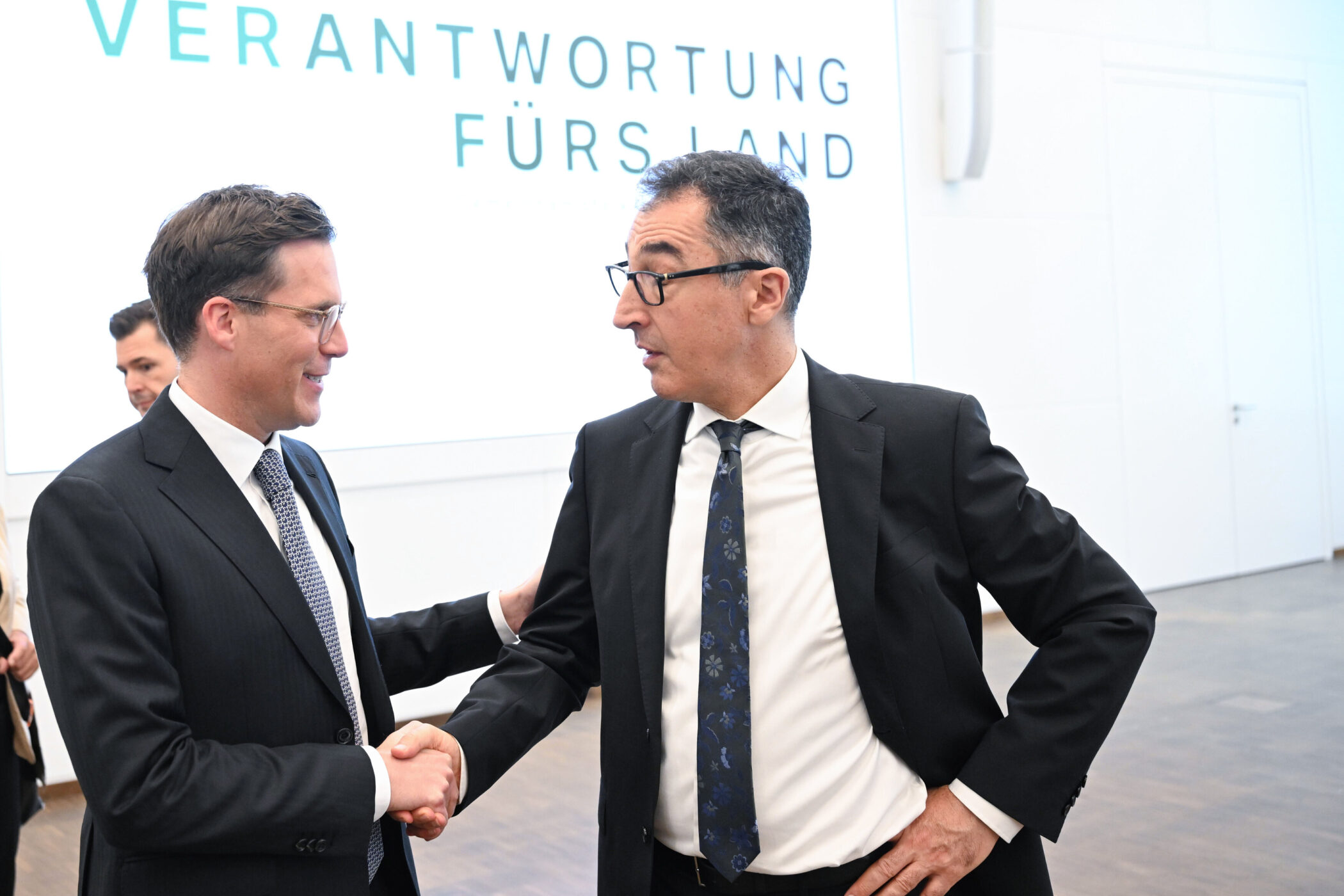 Zwei Männer im Anzug schütteln sich die Hände. Text im Hintergrund: „VERANTWORTUNG FÜRS LAND“.