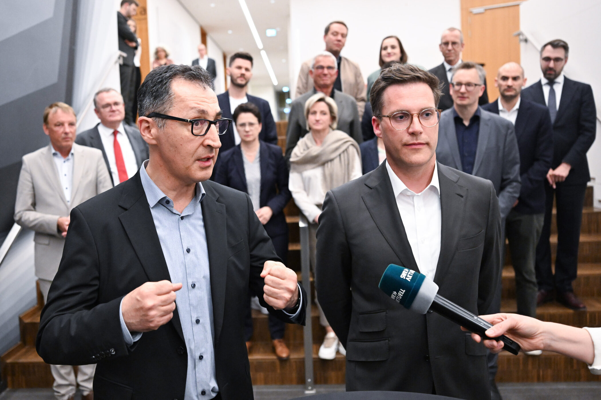 Zwei Männer im Anzug, einer spricht in ein Mikrofon. Mehrere Personen im Hintergrund.