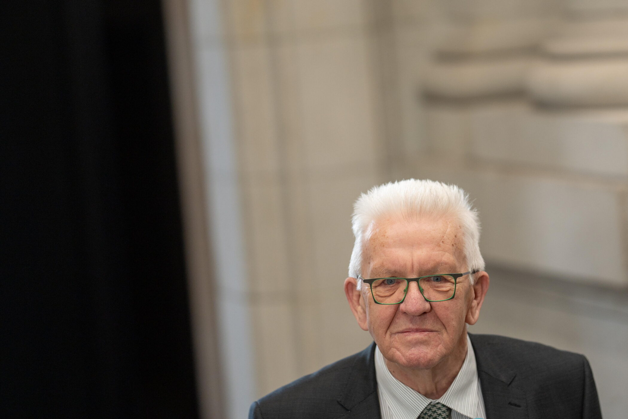 Älterer Mann mit Brille und Anzug vor unscharfem Hintergrund.