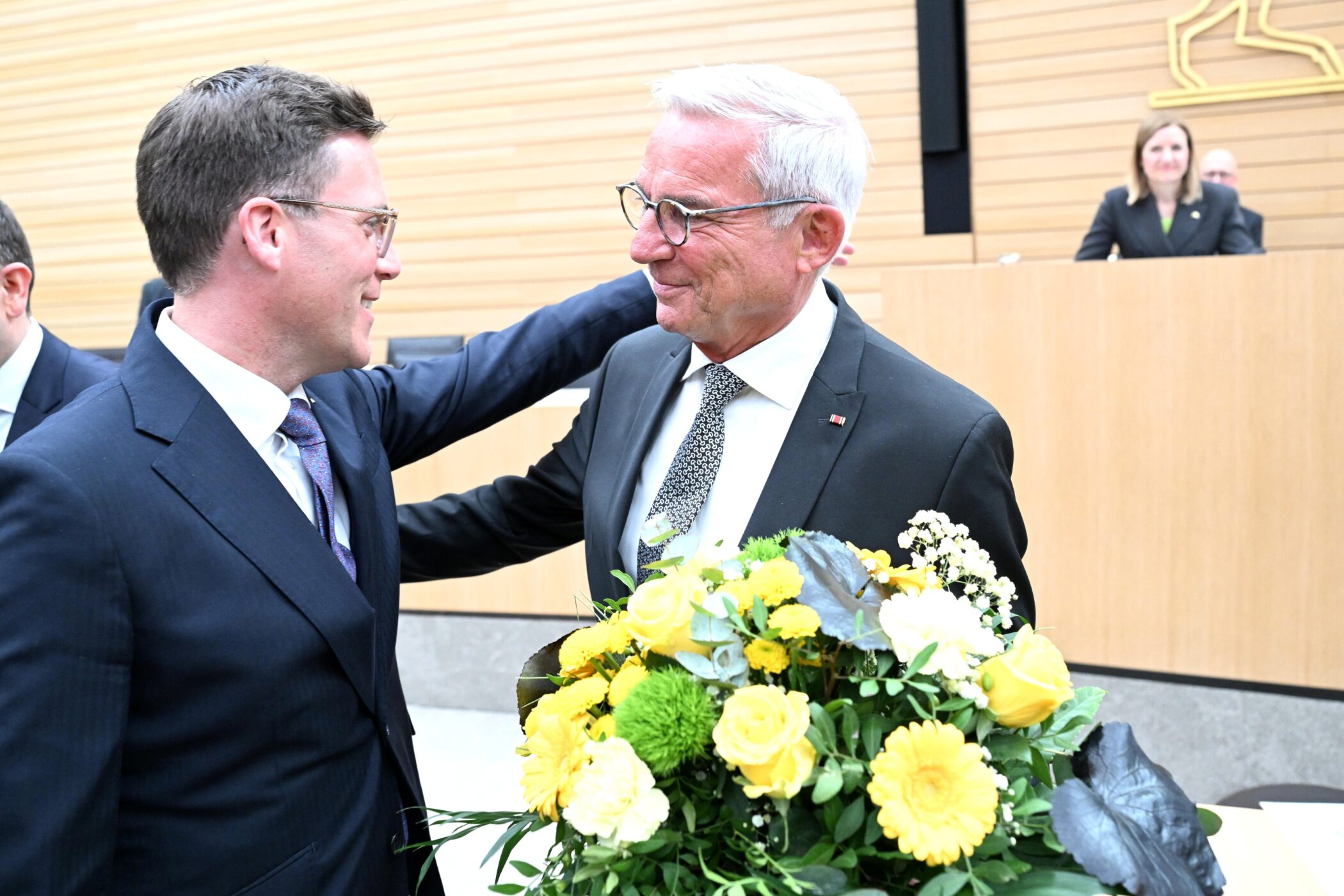 Zwei Männer im Anzug, einer hält einen Blumenstrauß, im Hintergrund zwei Personen.