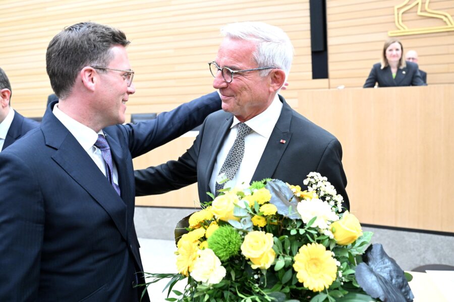 Zwei Männer im Anzug, einer hält einen Blumenstrauß, im Hintergrund zwei Personen.