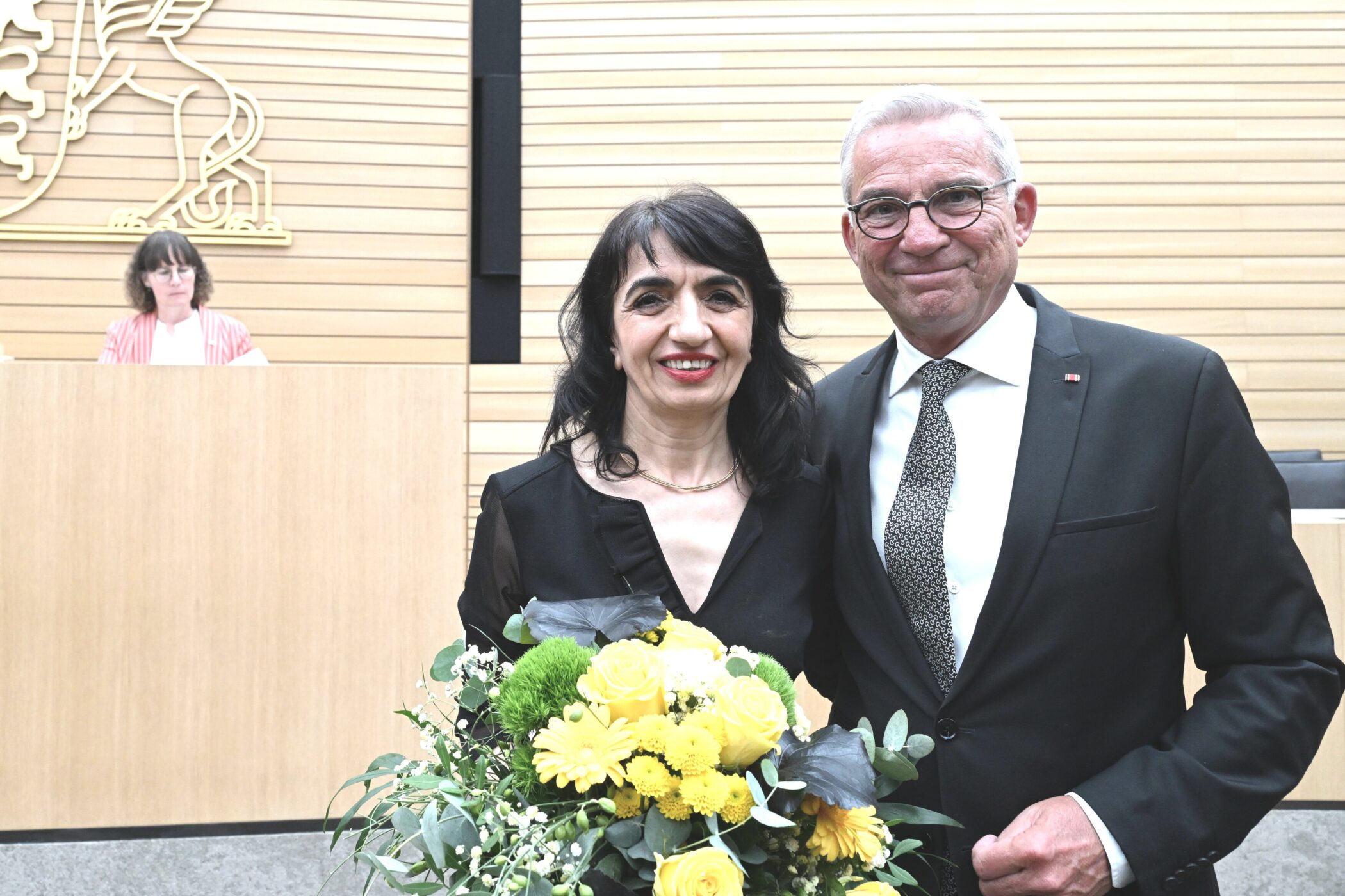 Zwei Personen mit Blumenstrauß, eine Frau sitzt im Hintergrund am Tisch.