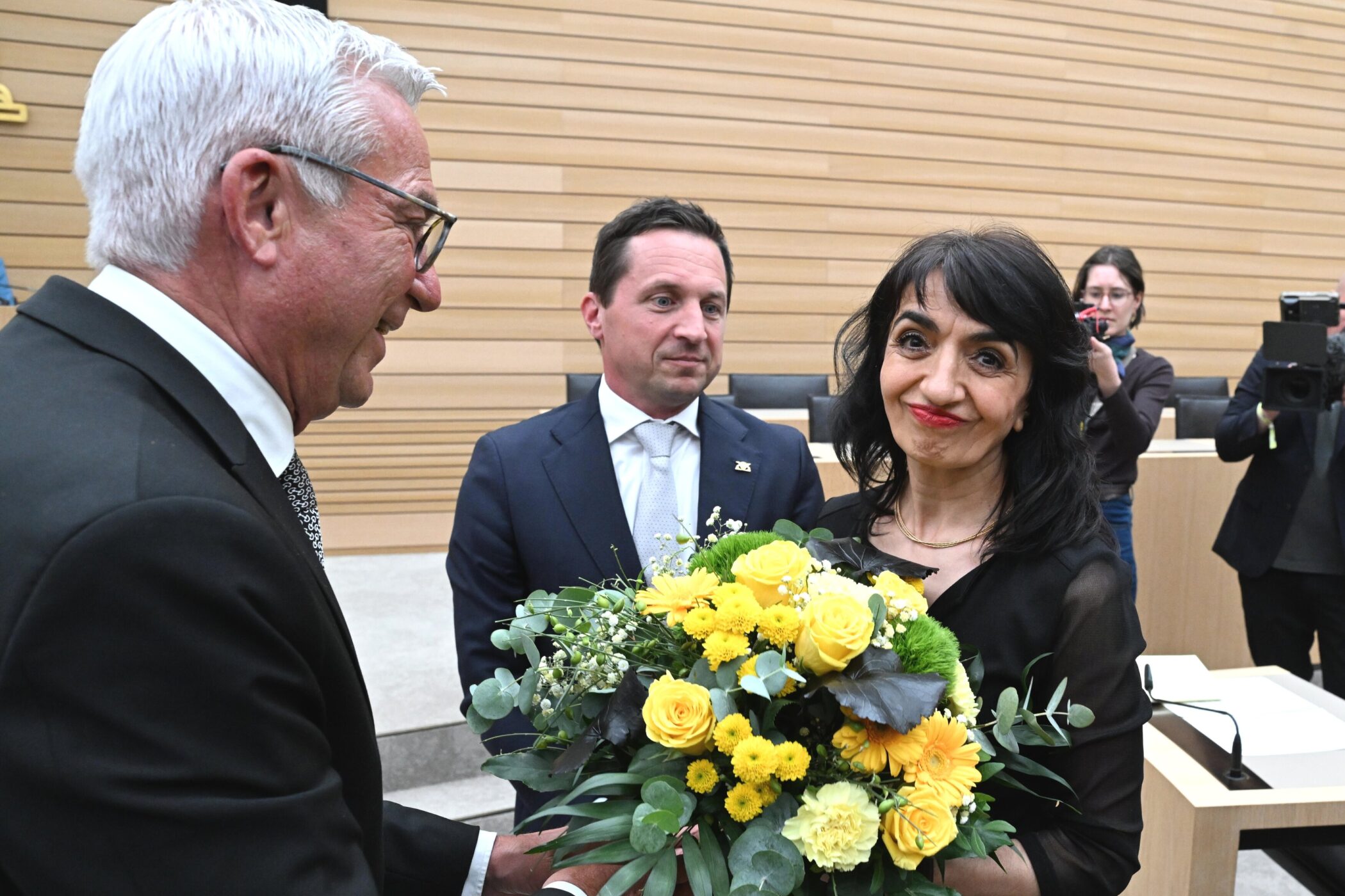 Drei Personen, eine hält einen Blumenstrauß. Fotografen im Hintergrund.