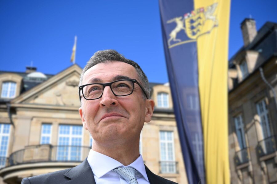 Mann mit Brille vor historischem Gebäude, Flagge mit Wappen im Hintergrund.