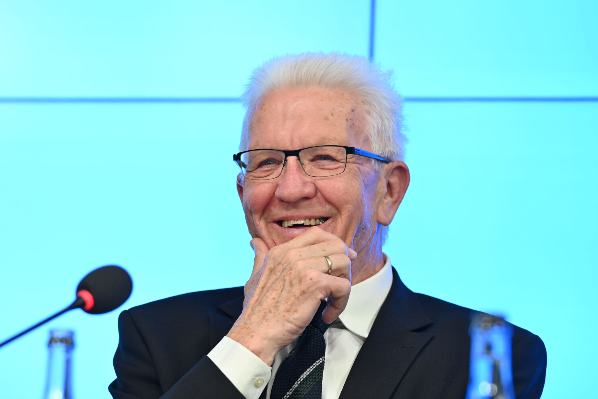 Älterer Mann mit Brille lächelt, hält seine Hand am Kinn, Mikrofon links.