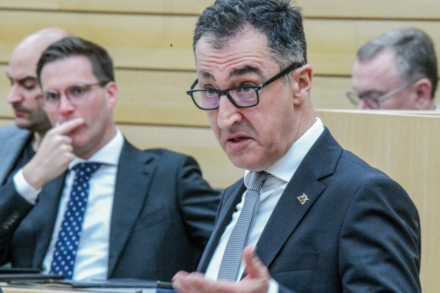 Drei Männer in Anzügen sitzen in einem Meetingraum. Einer spricht.